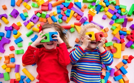 Zwei Kinder halten sich Spielholzklötze vor die Augen und gucken in die Kamera. Um sie herum liegen viele bunte Bauklötze Zwei Kinder halten sich Spielholzklötze vor die Augen und gucken in die Kamera. Um sie herum liegen viele bunte Bauklötze