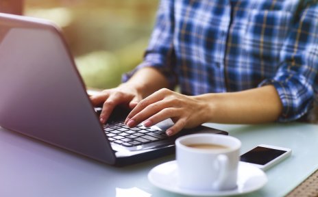 Eine Frau tippt auf einer Laptoptastatur, daneben Kaffeetasse und Smartphone. Eine Frau tippt auf einer Laptoptastatur, daneben Kaffeetasse und Smartphone.
