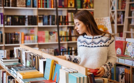 Eine junge Frau steht in einer Buchhandlung vor einem tischhohen Regal und schaut sich die Bücher an. Im Hintergrund stehen große Bücherregale. Eine junge Frau steht in einer Buchhandlung vor einem tischhohen Regal und schaut sich die Bücher an. Im Hintergrund stehen große Bücherregale.