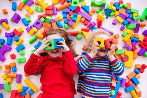 Zwei Kinder halten sich Spielholzklötze vor die Augen und gucken in die Kamera. Um sie herum liegen viele bunte Bauklötze Zwei Kinder halten sich Spielholzklötze vor die Augen und gucken in die Kamera. Um sie herum liegen viele bunte Bauklötze