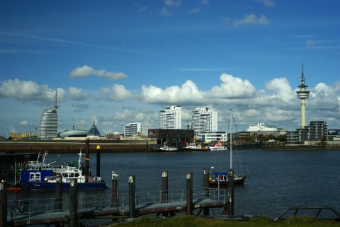 Bremerhavens Skyline ist in Form von Gebäuden und einem Turm im Hintergrund zu sehen. Davor ist ein Wasserkanal zu sehen. Im Vordergrund führt ein Steg zum Wasser. Einige Schiffe sind links im Bild festgemacht. Bremerhavens Skyline ist in Form von Gebäuden und einem Turm im Hintergrund zu sehen. Davor ist ein Wasserkanal zu sehen. Im Vordergrund führt ein Steg zum Wasser. Einige Schiffe sind links im Bild festgemacht.