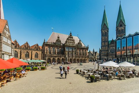 Aufnahme eines Platzes, auf dem Tische, Stühle und Sonnenschirme stehen. Der Platz ist von mehreren Gebäuden umgeben. Aufnahme eines Platzes, auf dem Tische, Stühle und Sonnenschirme stehen. Der Platz ist von mehreren Gebäuden umgeben.