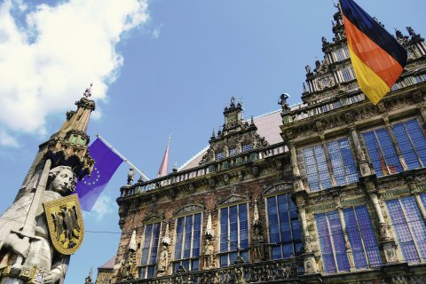 Froschperspektive auf eine steinerne Statue, die ein Schild und ein Schwert hält und auf ein Gebäude, an dem die Flagge Deutschlands und Europas hängt. Froschperspektive auf eine steinerne Statue, die ein Schild und ein Schwert hält und auf ein Gebäude, an dem die Flagge Deutschlands und Europas hängt.
