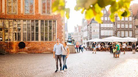 Eine Frau und ein Mann laufen am Bremer Rathaus vorbei. Im Hintergrund sieht man mehrere Menschen bei den Bremer Stadtmusikanten stehen. Eine Frau und ein Mann laufen am Bremer Rathaus vorbei. Im Hintergrund sieht man mehrere Menschen bei den Bremer Stadtmusikanten stehen.